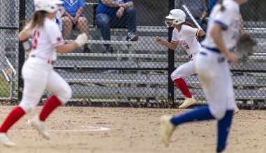 Two softball players in red and white uniforms run the bases with an opposing player in white and blue in the foreground
