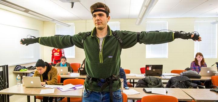 Student wearing motion-capture suit stands with arms out while classmates work in the background