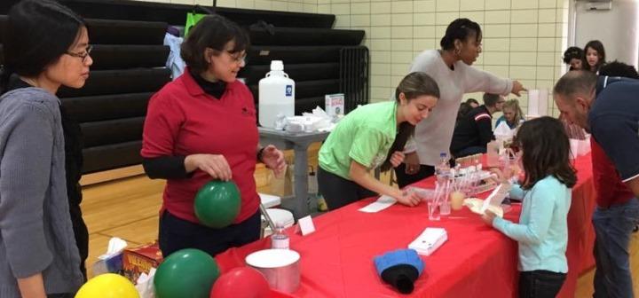Faculty hold balloons for an experiment while college students assist community members with the acid-base test. 