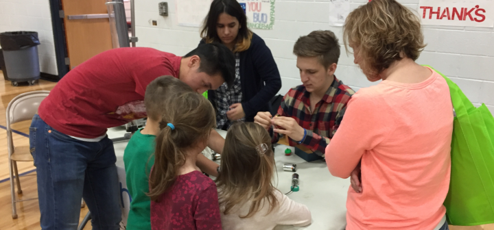 Children and college students huddle around a table where electronic circuits are constructed.