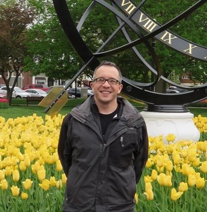 Nathaniel Rosi '99 in front of a large sundial and yellow tulips in Pella, IA.
