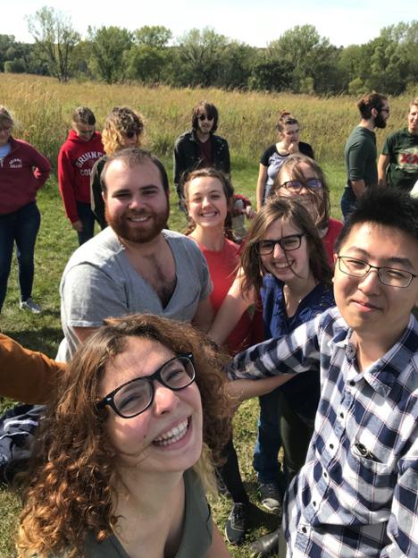 7 Grinnell Singers members stand in the background next to prairie grasses as 6 singers look joyously into the camera, appearing to be doing a team hand stack