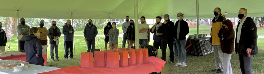 Chemistry graduates and faculty outside under a large tent, with gift bags and cupcakes to go on a covered table. 
