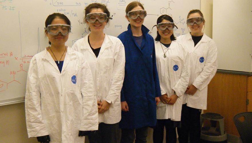 Four research students with their faculty mentor, Prof. Key, wearing lab coats and safety goggles in front of a markerboard showing chemical reactions