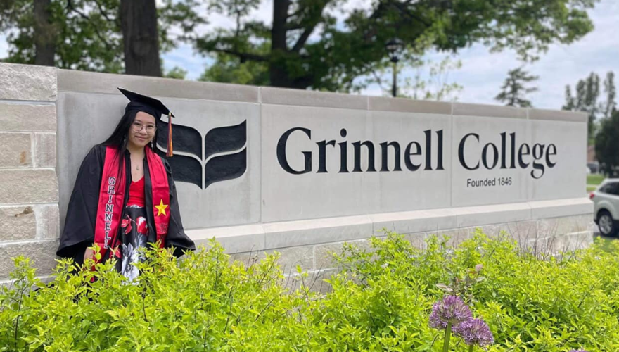 Nhi Vo poses in cap and gown near the Grinnell College sign, surrounded by greenery.