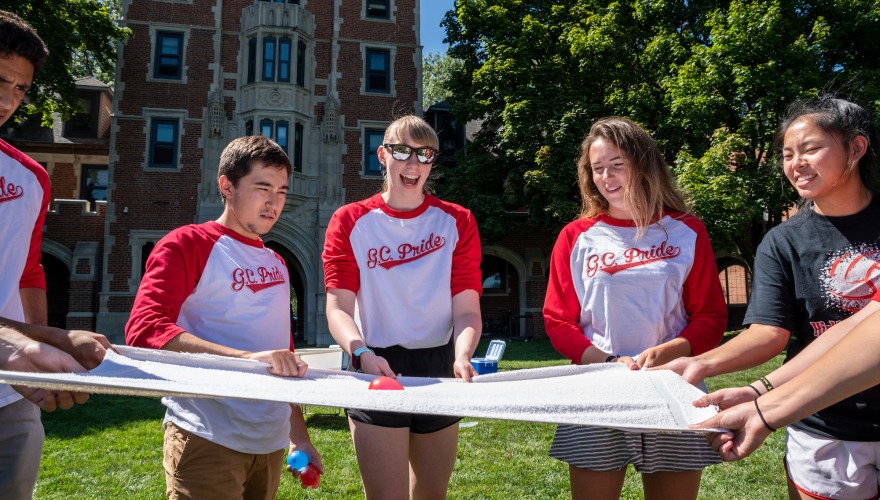 Students in Grinnell College shirts gathered around a sheet with a red object, smiling while playing a game
