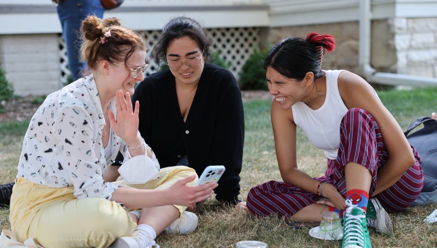 Three students sit on the grass and laugh while one of them shares something displayed on her phone.
