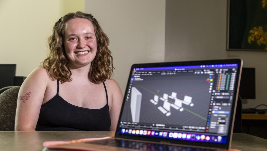 Kailee sits behind her computer. The screen faces the camera and shows a 3D model of a classroom.