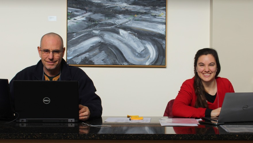 A man on the left and a woman on the right smile at the camera. They sit at a desk with their computers.