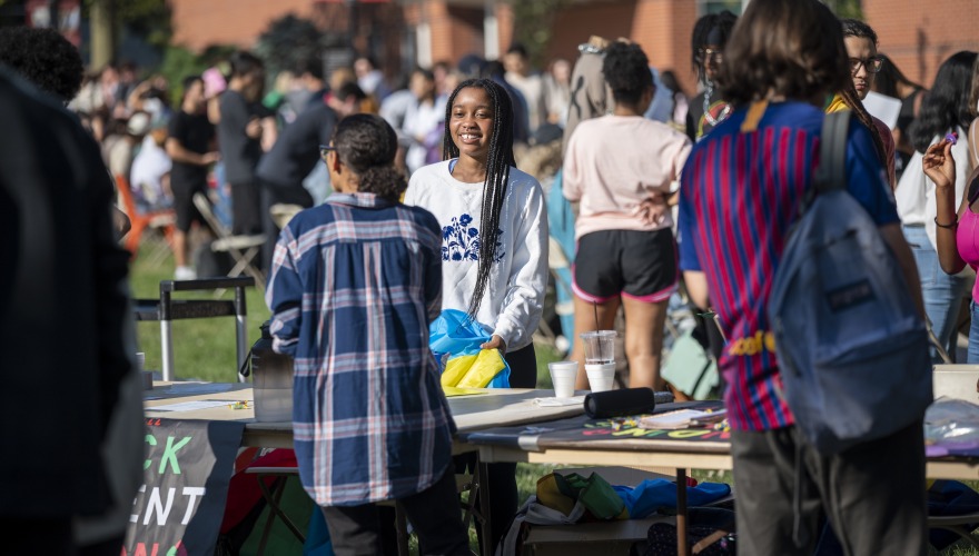 A Black student with braids smiles to another student. They are surrounded by many other students who are participating in the student organization fair.