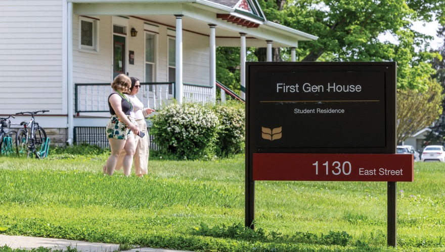 A large sign in front of a house reads in white letters on black First Gen House Student Residence the address, 1103 East, appears in white text on a red bar below. 2 women stroll by in the background