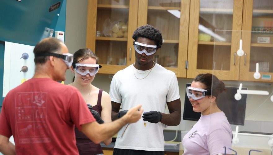 Three students wearing safety goggles listen to a chemistry professor explain a laboratory experiment.