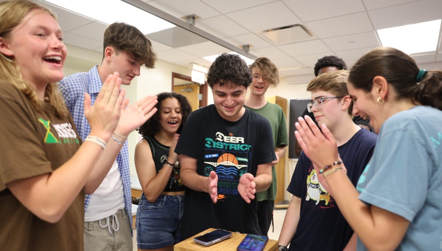 A group of students stands around a small wooden box clapping.
