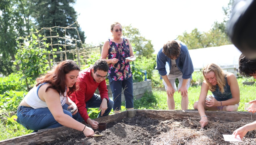 Four students crouch in front of a garden bed of soil under the supervision of a biology professor.