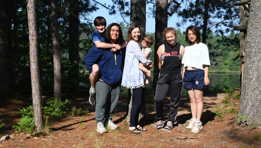 Four students pose with their professor, holding her baby, in the woods of Northern Michigan