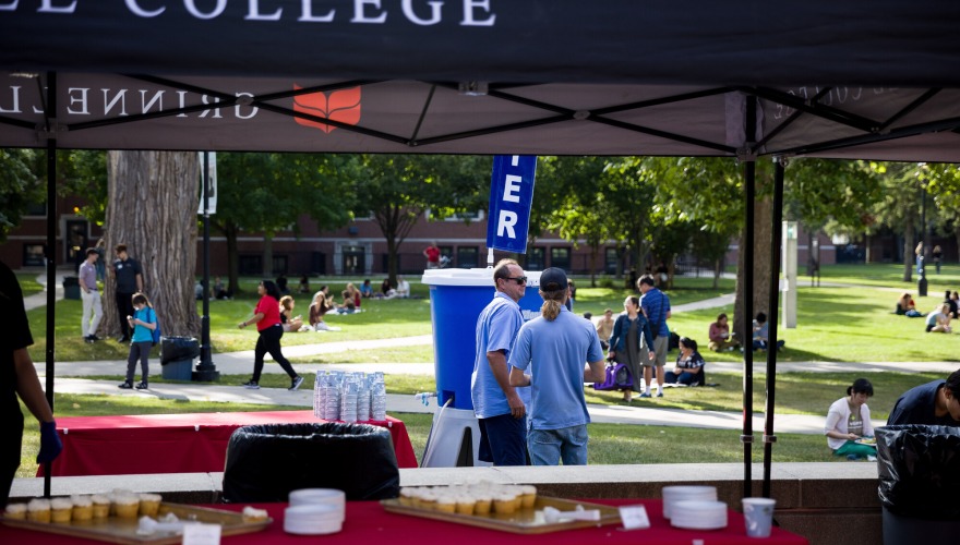 Two people wearing blue stand behind a food tent; many other people in the background sit in the grass eating.