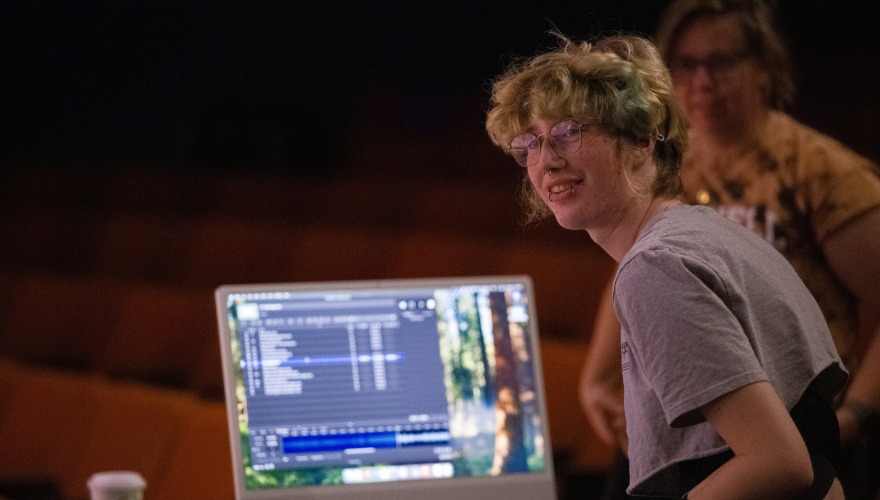 A student turned to face the camera with a monitor showing audio software in front of them