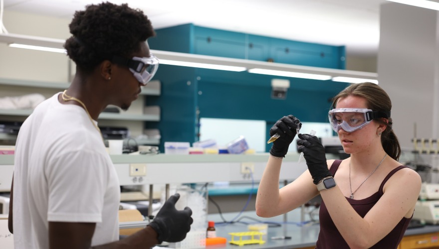 Two students wearing lab goggles and black rubber gloves study a pipet in a chemistry lab.
