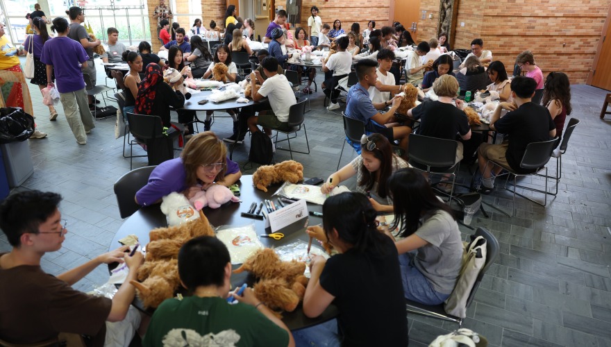 A large group of students sit at tables building stuffed animals.