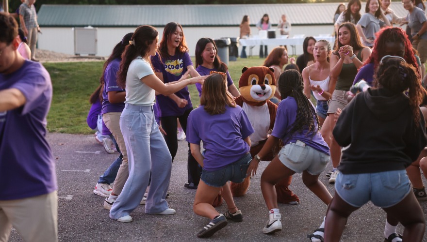 A group of students, mostly in purple shirts, dance with a squirrel mascot.