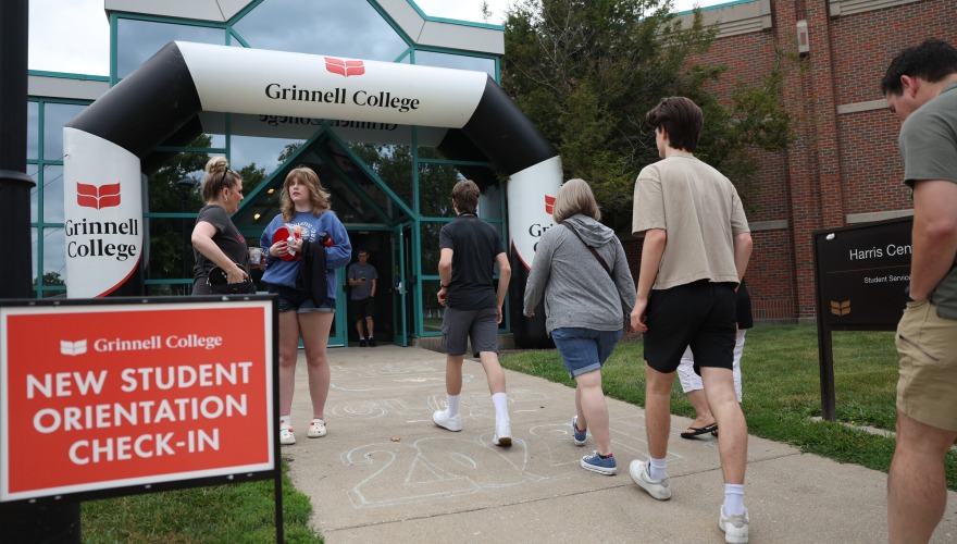 People walk into a building through a black and white inflatable arch that is branded with "Grinnell College". A red sign with white text reads "New Student Orientation Check-In"