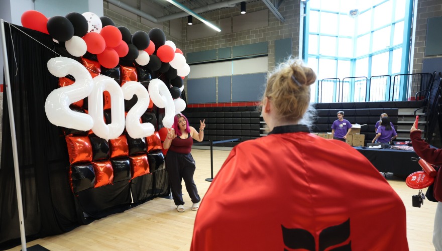 The back of a woman wearing a red cape with a black laurel leaf logo. She stands in front of a photo wall with red and black balloons and white balloons that read "2029"