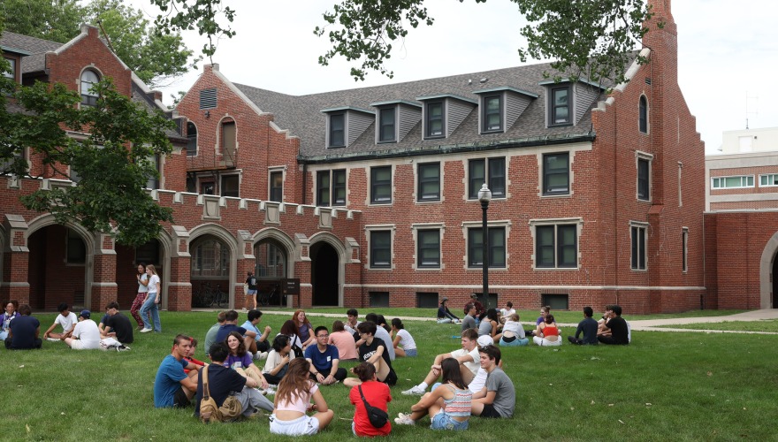 Students sit in circles on the grass with a brick building in the background.