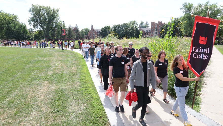 First-year students parade down a sidewalk with one at the beginning of the line holding a sign that reads "Grinnell College"