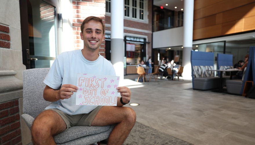 Student James Snyder sits in a chair and smiles while holding a sign that reads "First Day of School!"