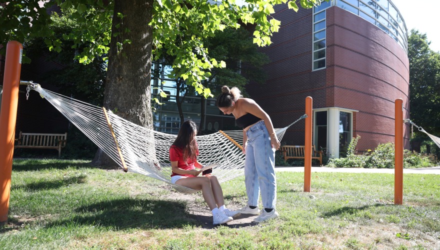 A student sitting in a hammock holding a piece of paper while another student stands beside and looks on.