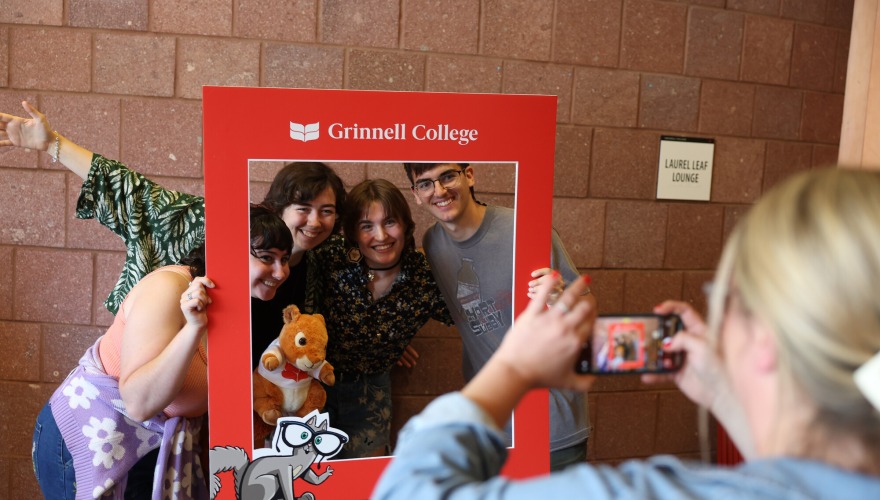 Four students pose behind a red photo frame while a woman takes their picture.