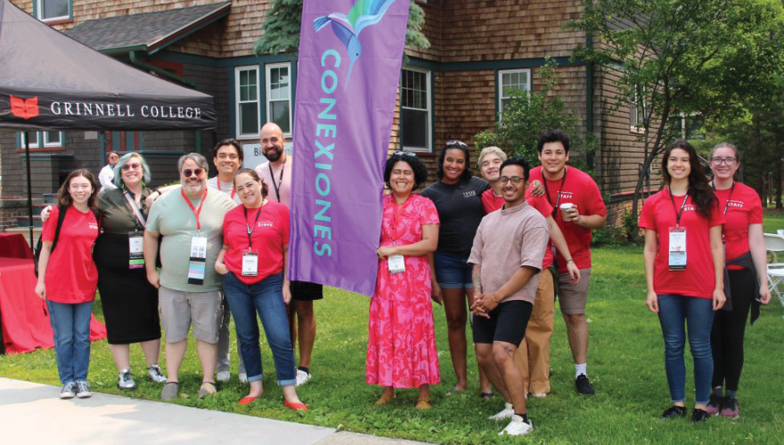 Alumni on campus gather around a tall purple sign with the word Conexiones in blue text with their bird icon in shades of blue above that.