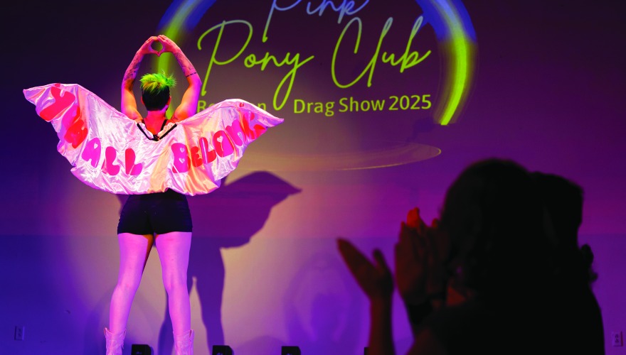 A person with back to audience and hands in a heart gesture wears wings that read You All Belong. Pony Club and drag show 2025 are displayed in light on the backdrop