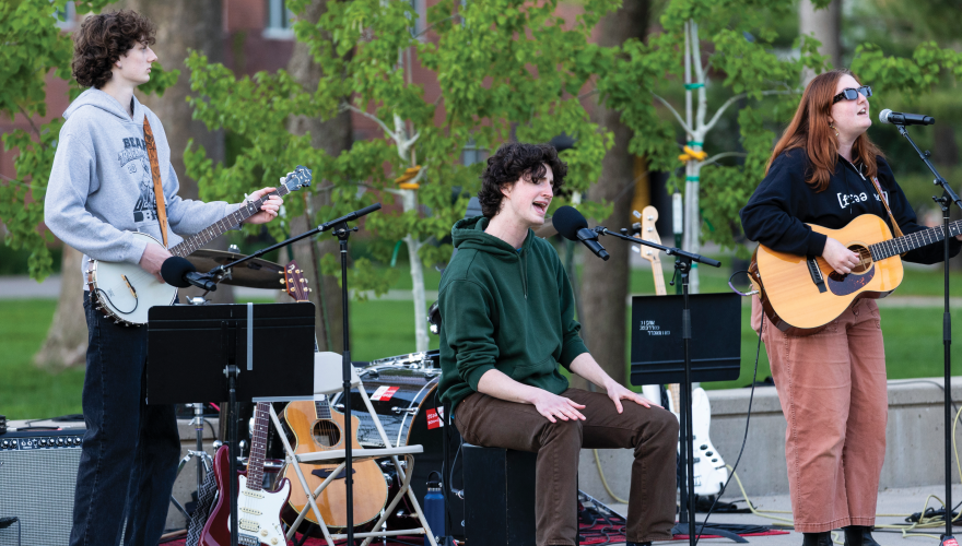 at an outdoor location, Ben Curran sings at a mic while two other students who are playing guitar and bass 