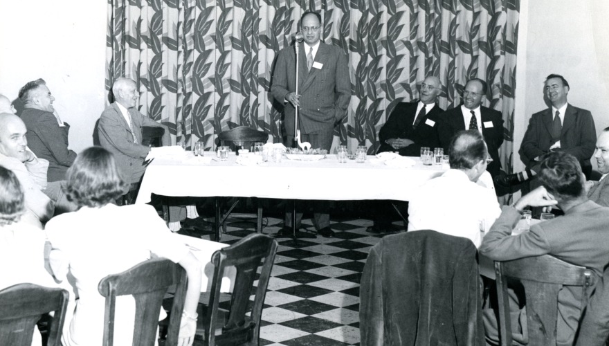 Black and white photo of a meeting with one man speaking at a podium