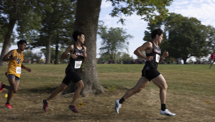 Three male college students wearing cross country uniforms and race bibs running one after another