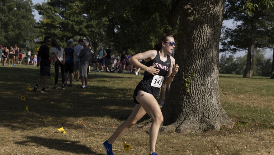 A female college student wearing a cross country uniform, race bib, and sunglasses running on a grassy course