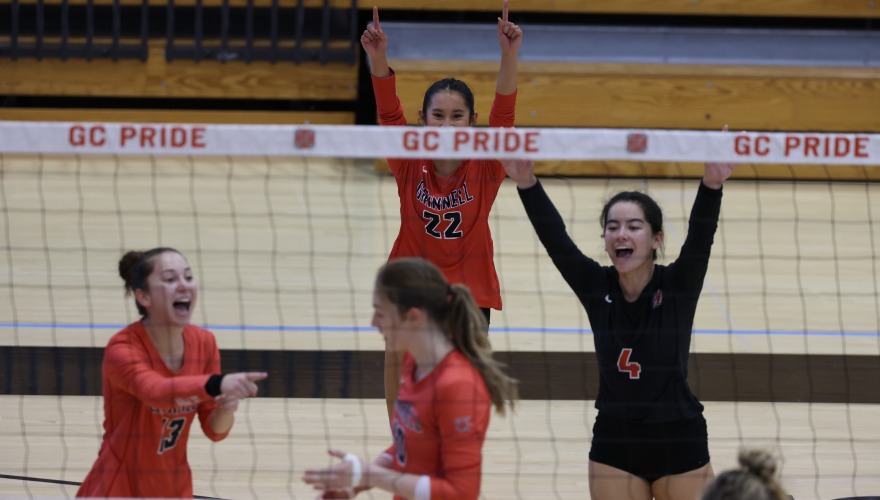 Four volleyball players in red and black uniforms celebrate on the far side of the net