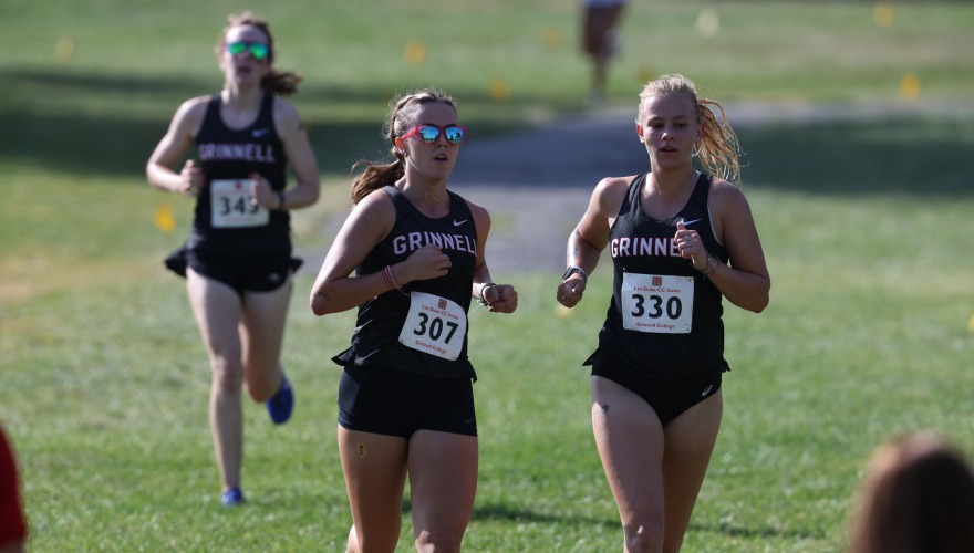 Two female college students wearing cross country uniforms run together, a third behind them
