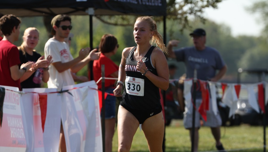 A female college student wearing a cross country uniform and race bib running as people clap on the sidelines