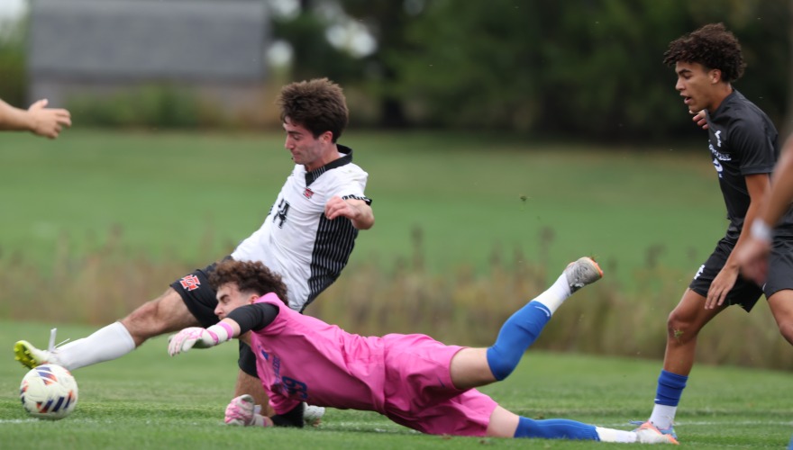 A soccer player wearing a white uniform with the ball at his foot while a goalie in pink dives for it