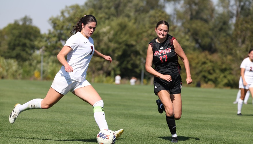 A women's soccer player wearing a white uniform about to kick the ball with a player in black running after