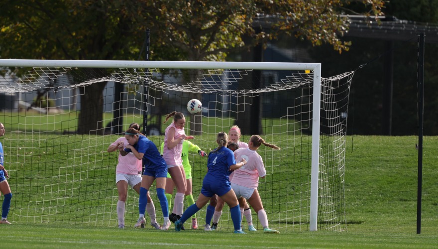 Women's soccer players in pink and blue crowd around the goal as one heads the ball out