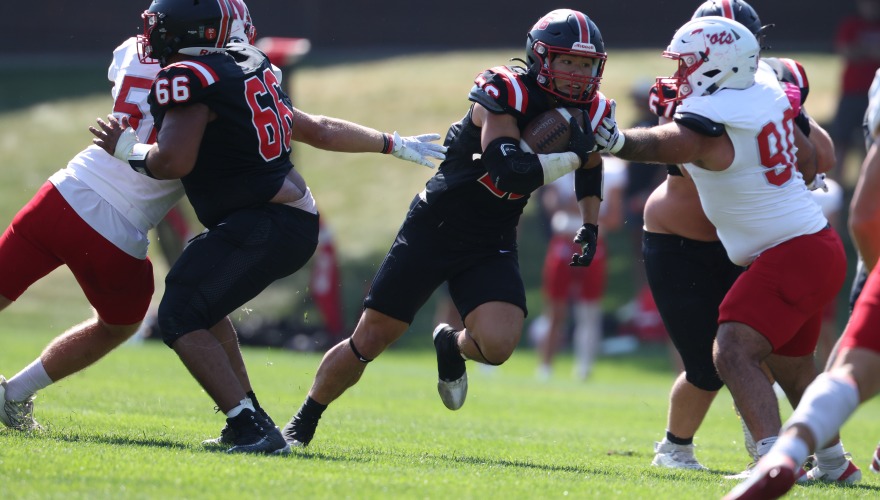 A football player in black runs the ball past defenders in white and red