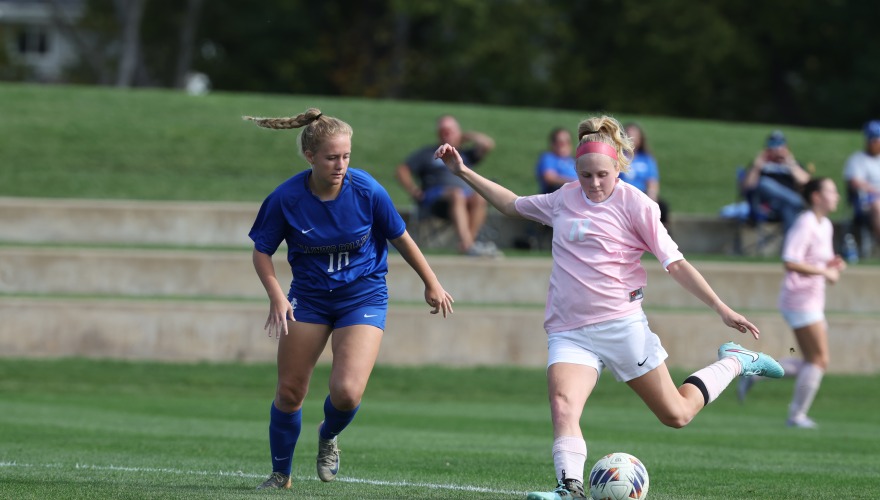 Grinnell women's soccer player in pink jersey about to kick the ball as a defender in blue approaches