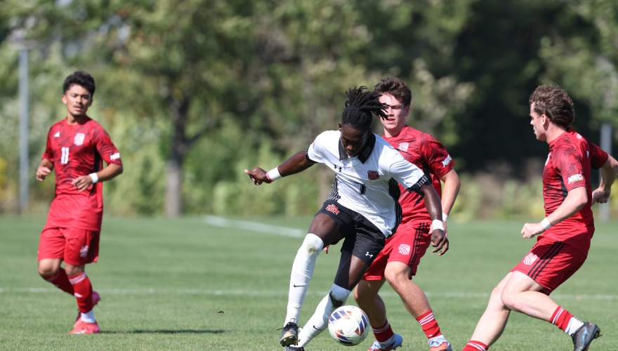 Grinnell men's soccer player in white runs with the ball at his feet around three players in red