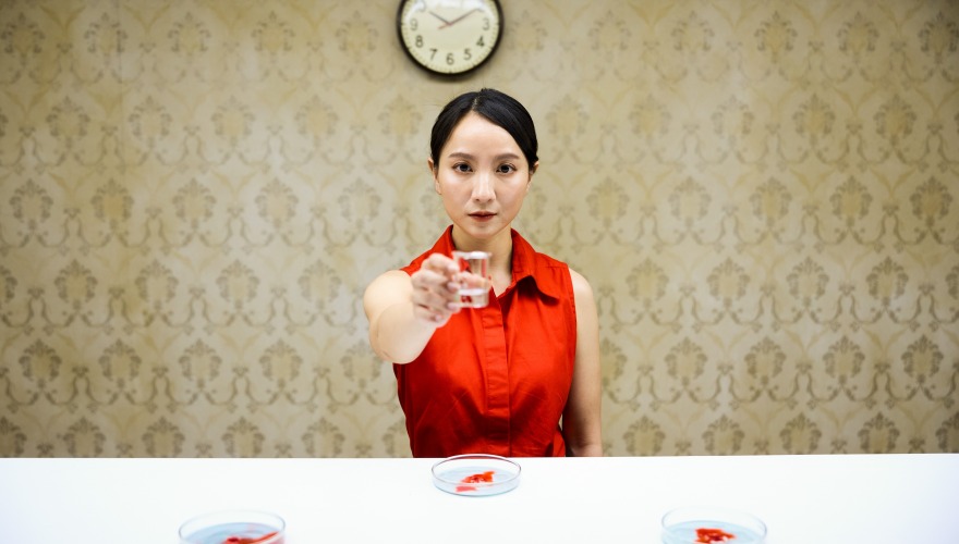 A young woman actress in a red blouse is handing a small glass to the viewer