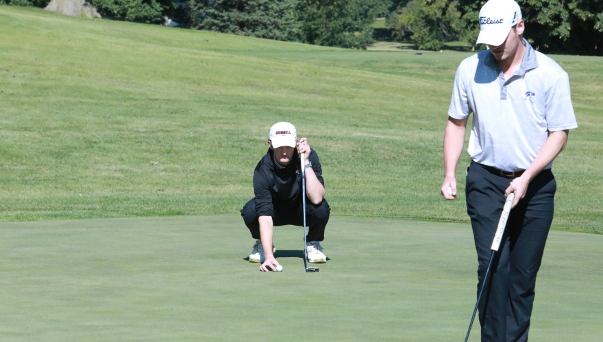 A golfer in black crouches lining up a putt with another in gray standing in the foreground