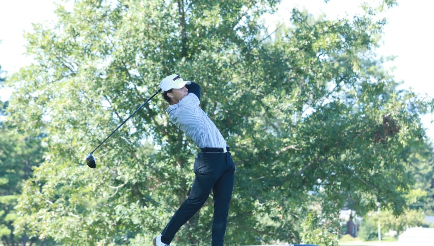 A golfer wearing a gray top, black pants, and white hat follows through after a shot