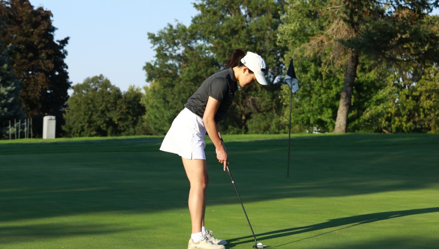 A golfer wearing a black top, white skirt, and white hat lines up a shot